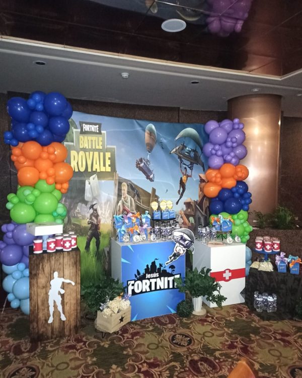 Imagen de decoración con globos de una primera comunión, se aprecia un panel de varios metros con una imagen gigante del Juego Fortnite con varias mesas con colores a juego delante y una guirnalda de globos a un lado
