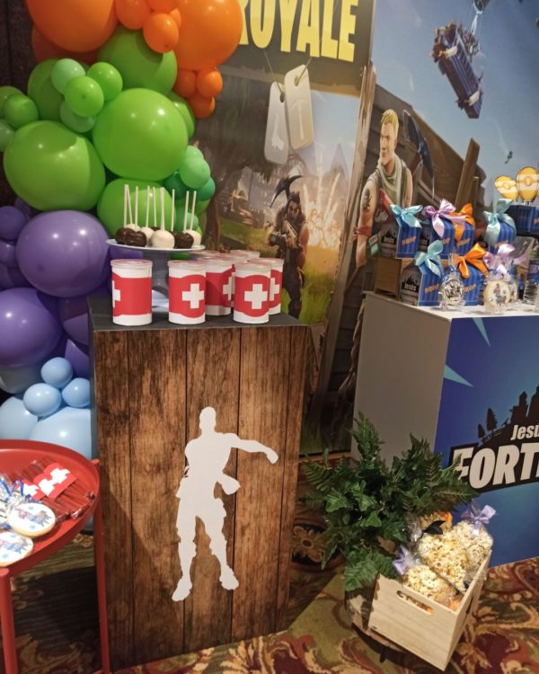 Imagen de detalle de la decoración con globos de una primera comunión, se aprecia un panel de varios metros con una imagen gigante del Juego Fortnite con varias mesas con colores a juego delante y una guirnalda de globos a un lado