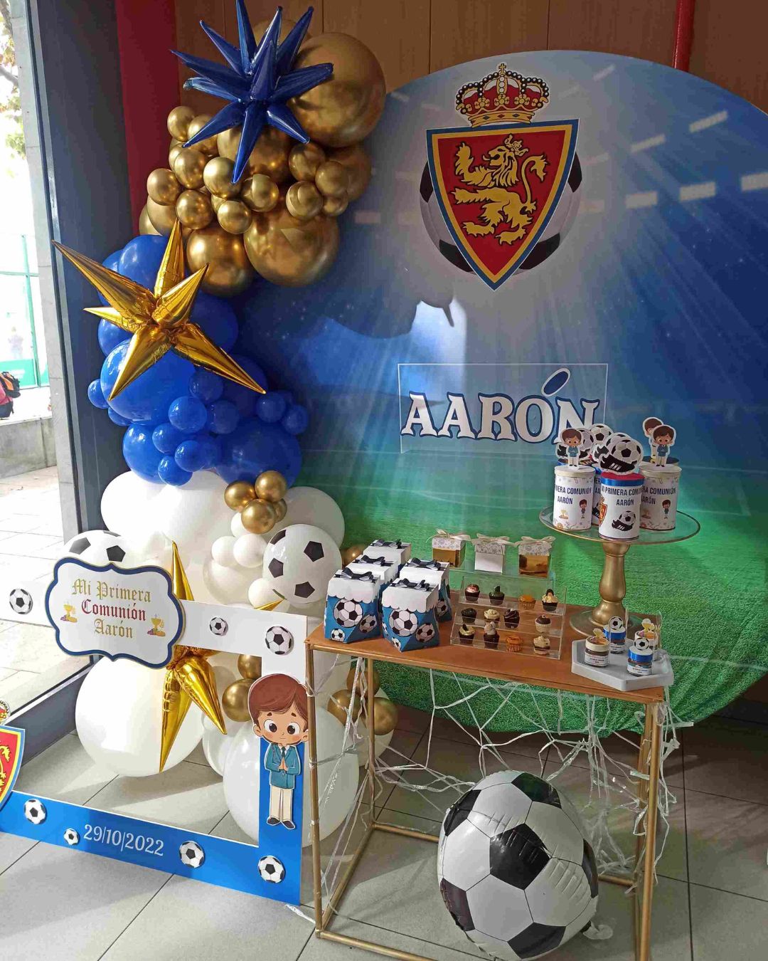 Decoración de comunión con motivo de Fútbol, con guirnalda en colores blanco azul y dorado y una trasera alegórica a un campo de fútbol