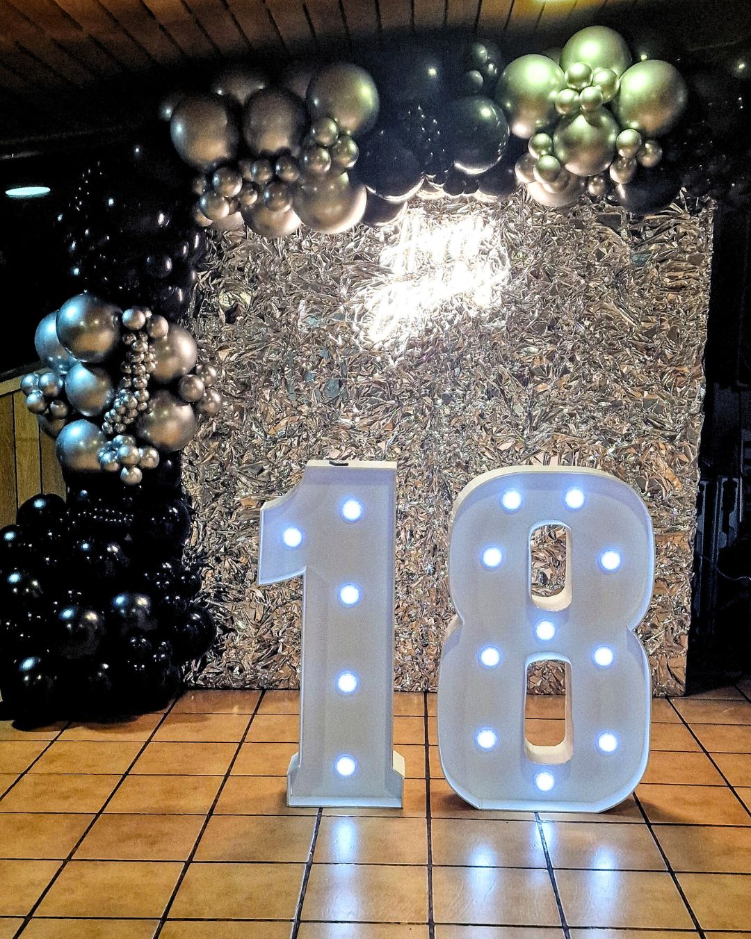Photocall Cumpleaños 18 años letras blancas con backing negro, guirnaldas de globos dorados, plata y negros