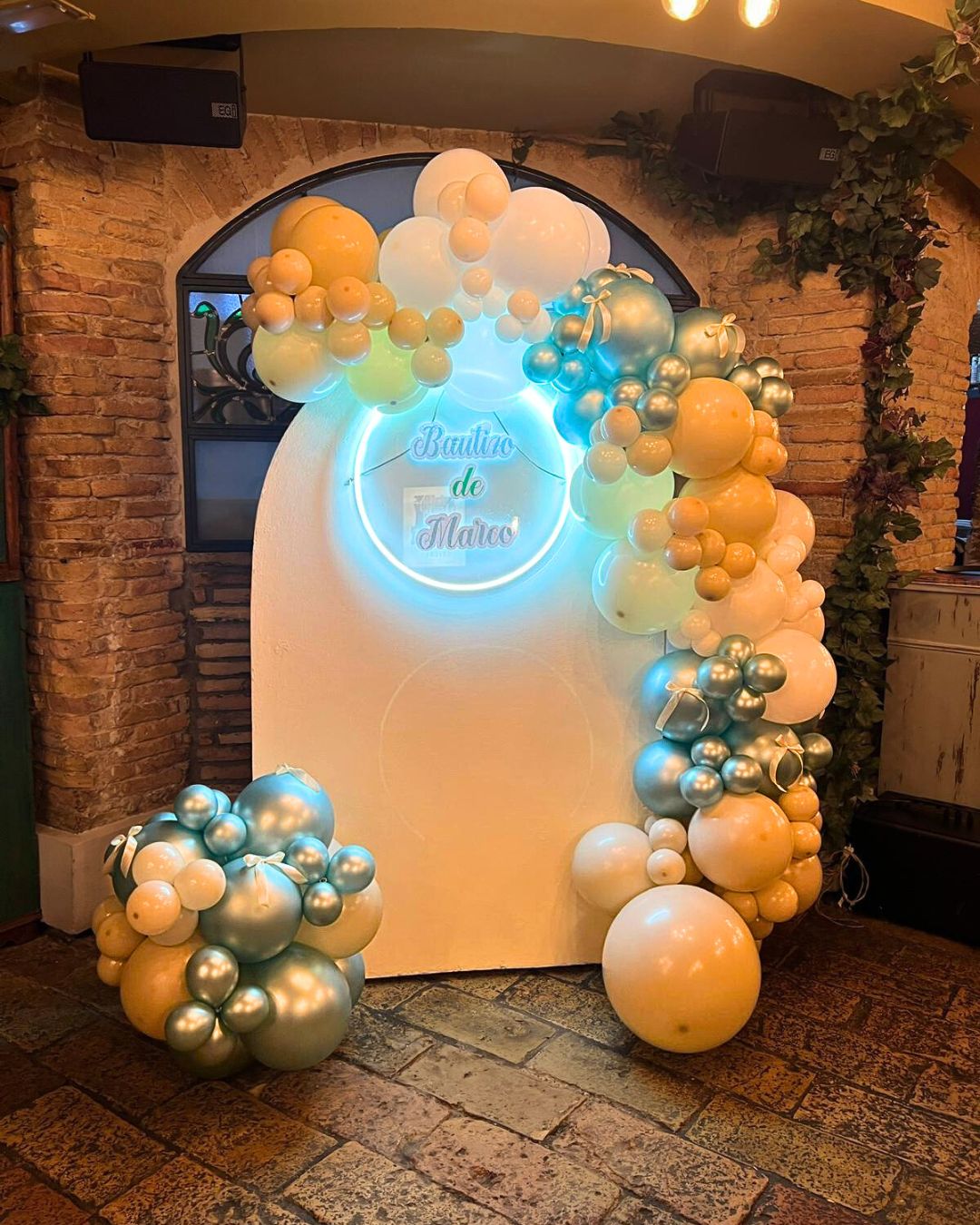 Panel color crema con guirnalda de globos de tonos azul pastel y a un lado del panel y cartel luminoso que dice Bautizo de Marco