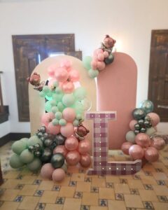 Panel en forma de arco con globos de colores pastel acompañado de otro panel rosado con mini guirnaldas de globos. y una letra L gigante de 1 metro de alto con textura y colores a juego