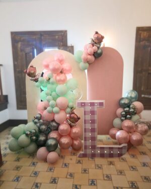 Panel en forma de arco con globos de colores pastel acompañado de otro panel rosado con mini guirnaldas de globos. y una letra L gigante de 1 metro de alto con textura y colores a juego